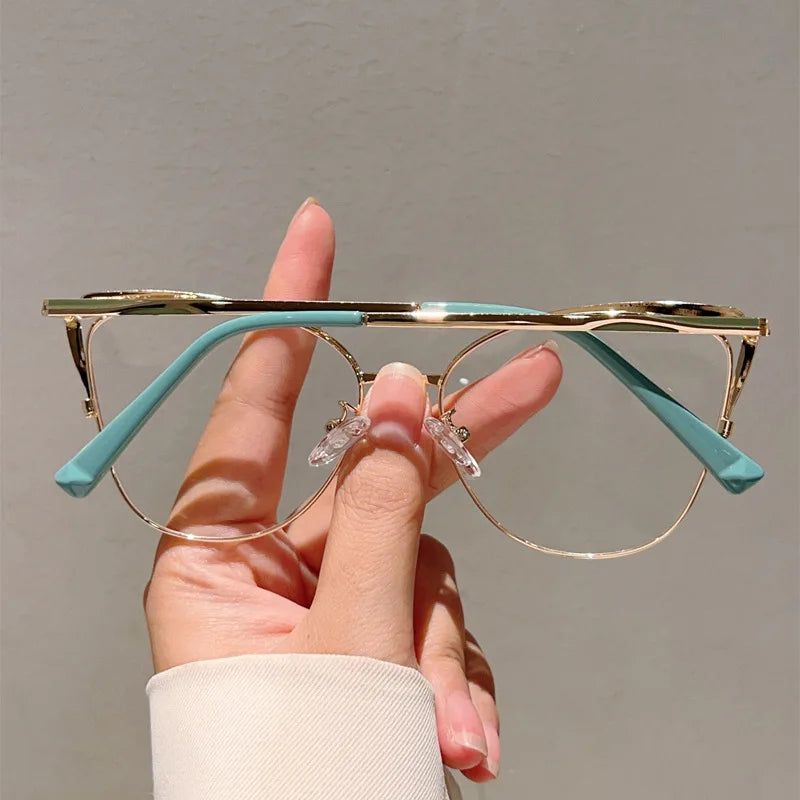 Una mano sostiene unas gafas de computadora estilo ojo de gato de metal azul claro con detalles dorados contra un fondo neutr