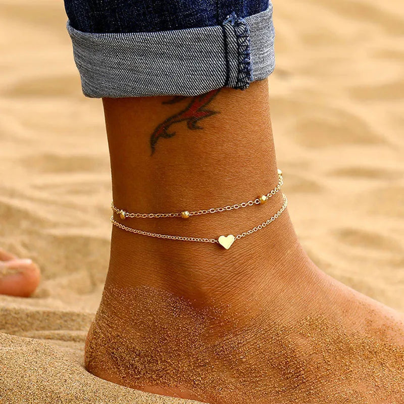 A close-up of a woman's ankle showing an elegant geometric anklet bracelet on her skin against a sandy background.