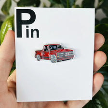 A red pickup truck metal clip pin is displayed on a white card against a green leafy background.