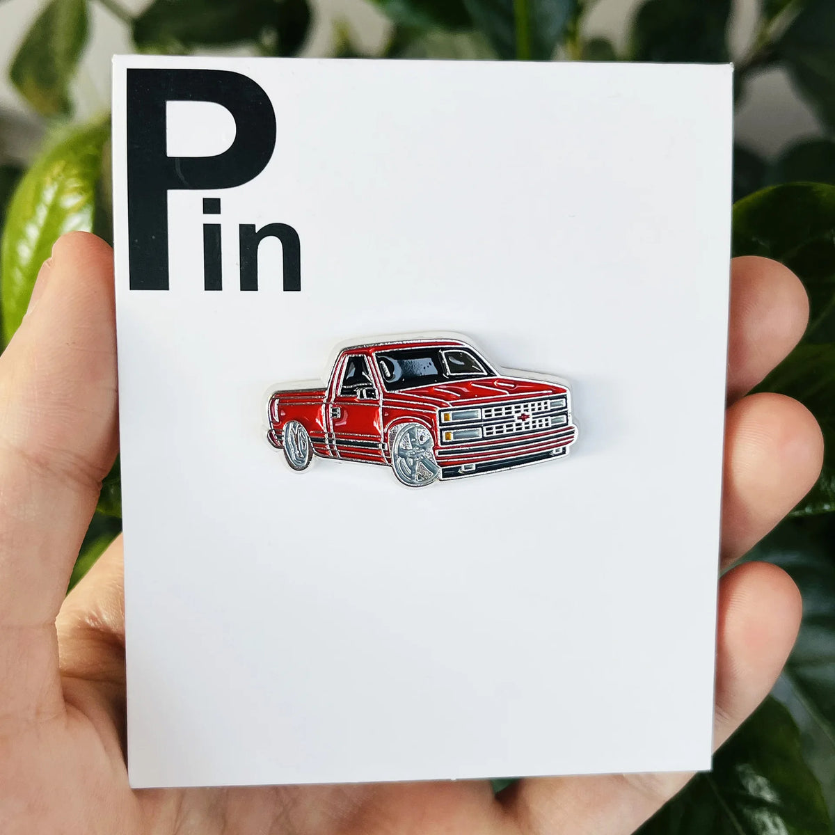 A red pickup truck metal clip pin is displayed on a white card against a green leafy background.