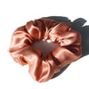 A close-up of a satin rose gold hair scrunchie, rolled and draped, casting a shadow on a white surface.