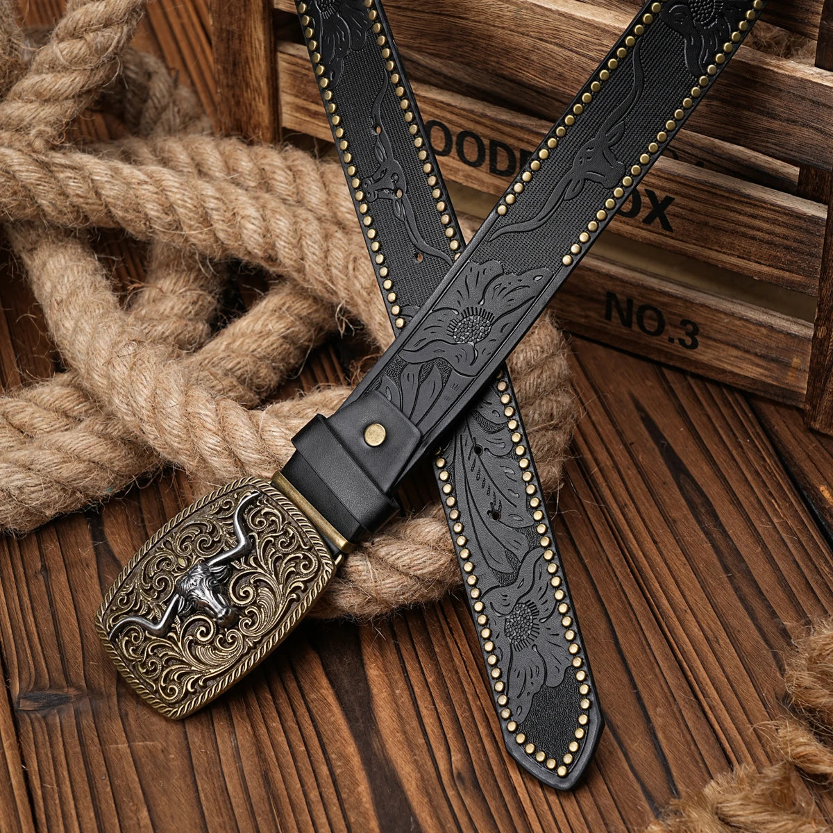 A black PU leather belt with a floral engraving and brass buckle is laid across a thick rope on a wooden surface.