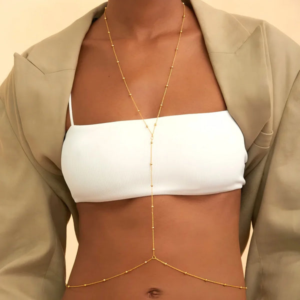 A close-up of a model wearing a gold adjustable belly chain body jewelry over a white crop top and beige jacket.