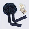 A dark navy silk sleep bonnet with a gathered top and long ties, next to a bowl of white hair accessories.