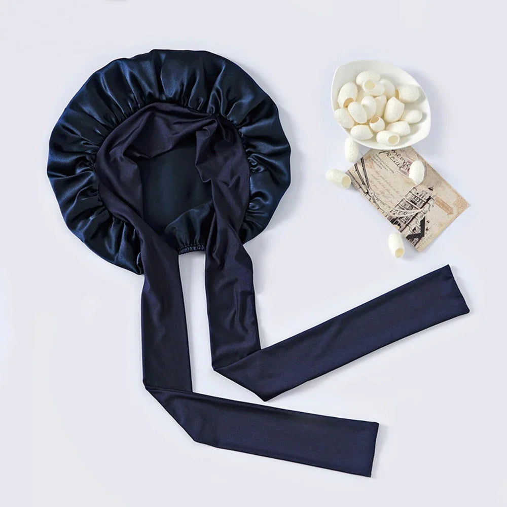 A dark navy silk sleep bonnet with a gathered top and long ties, next to a bowl of white hair accessories.