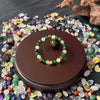 A natural stone and wooden bead prosperity bracelet rests on a dark brown circular stand surrounded by various colorful polis