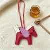 A red horse pendant charm with purple wings is displayed on a cream-colored fabric next to a small glass jar of honey.