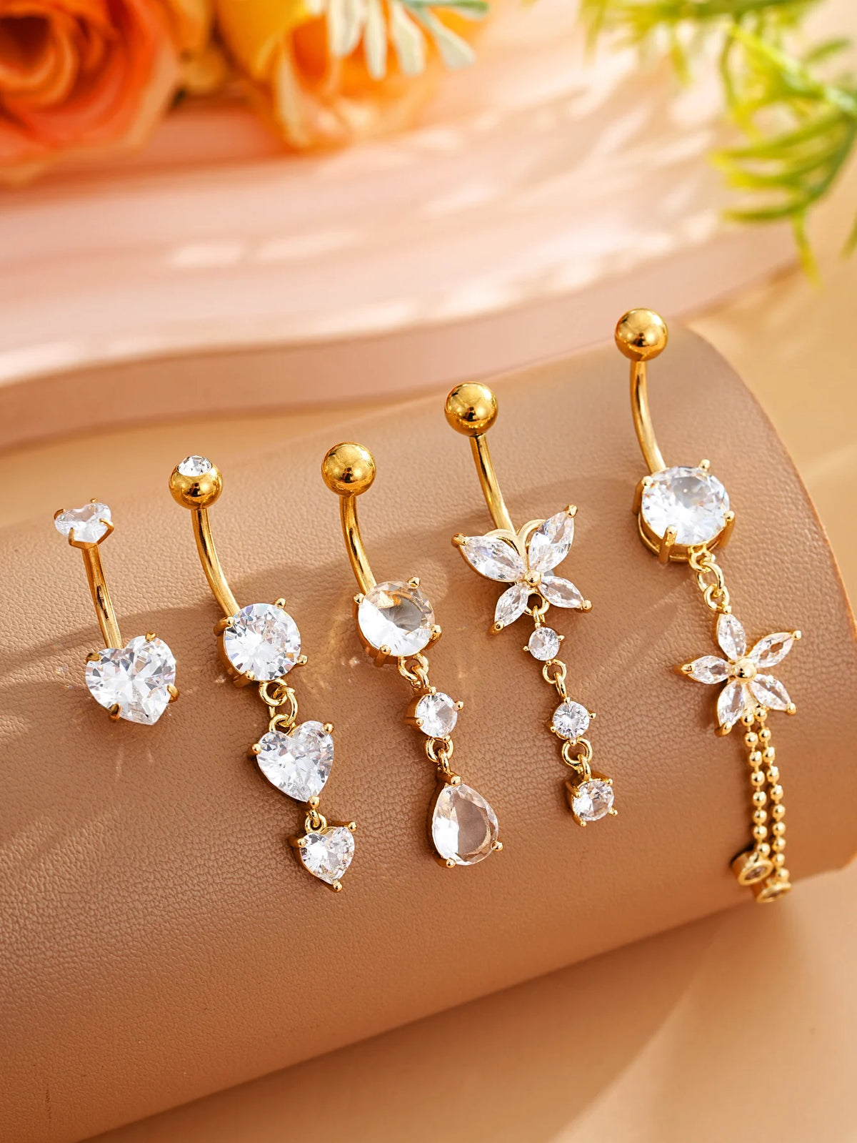 A set of five gold stainless steel dangled belly button rings with clear and floral charms displayed on a beige surface.