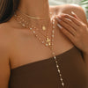 A close-up of a woman wearing a layered gold necklace with pearl beads and a cross pendant against her bare shoulder.