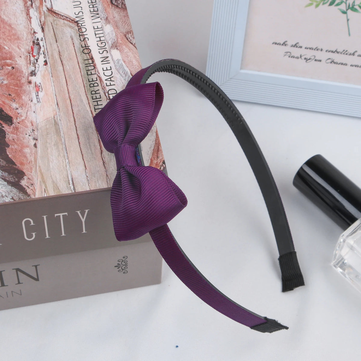 A purple grosgrain ribbon hair bow is displayed next to a book and a black cosmetic product.