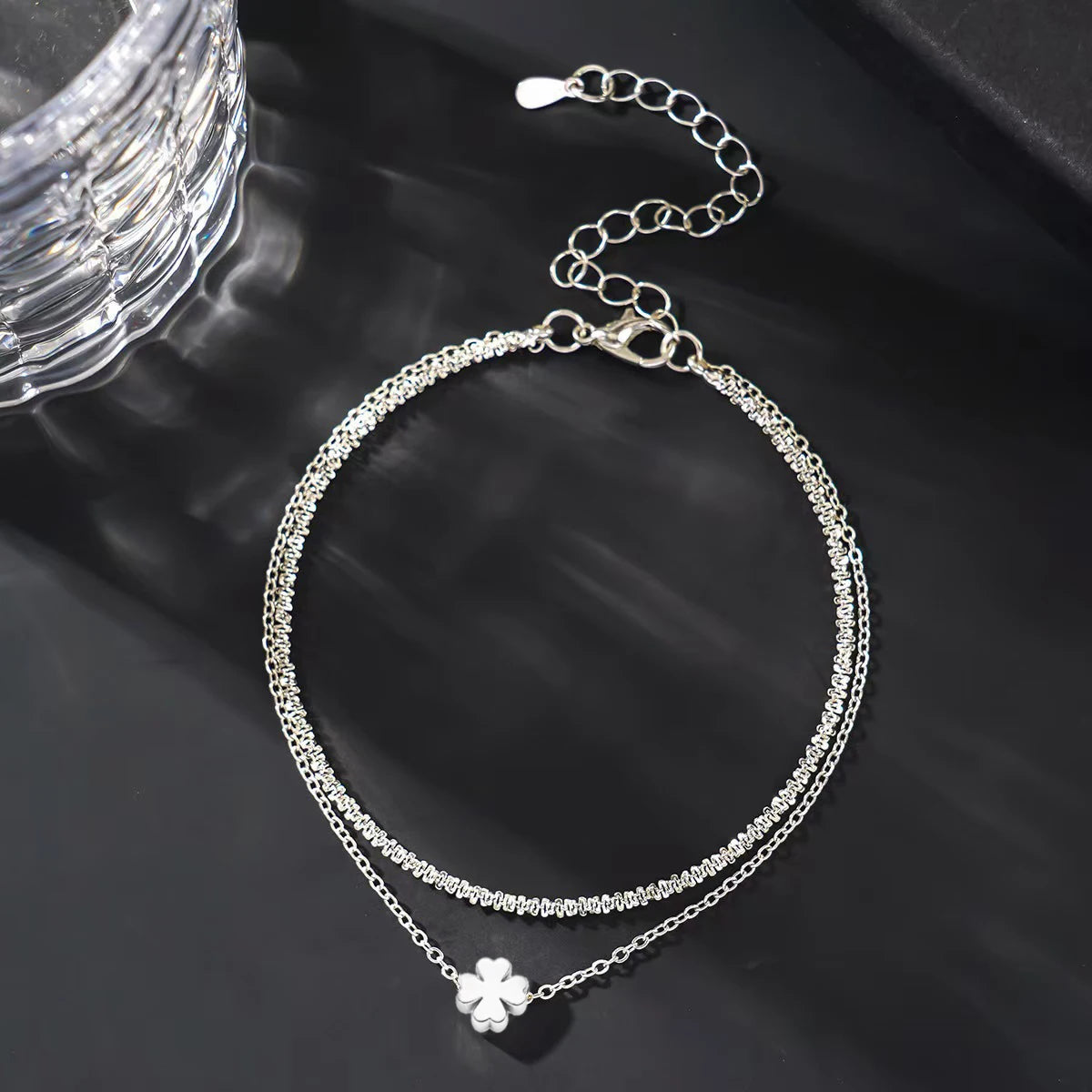 A delicate silver-plated chain anklet with a small flower charm displayed on a dark surface.