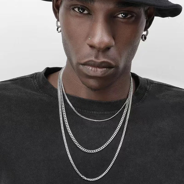 A close-up of a man wearing a layered stainless steel Cuban chain necklace against a dark shirt.