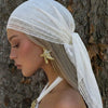 A model wearing a white bohemian lace trim shawl draped over her head and shoulder against a tree trunk.