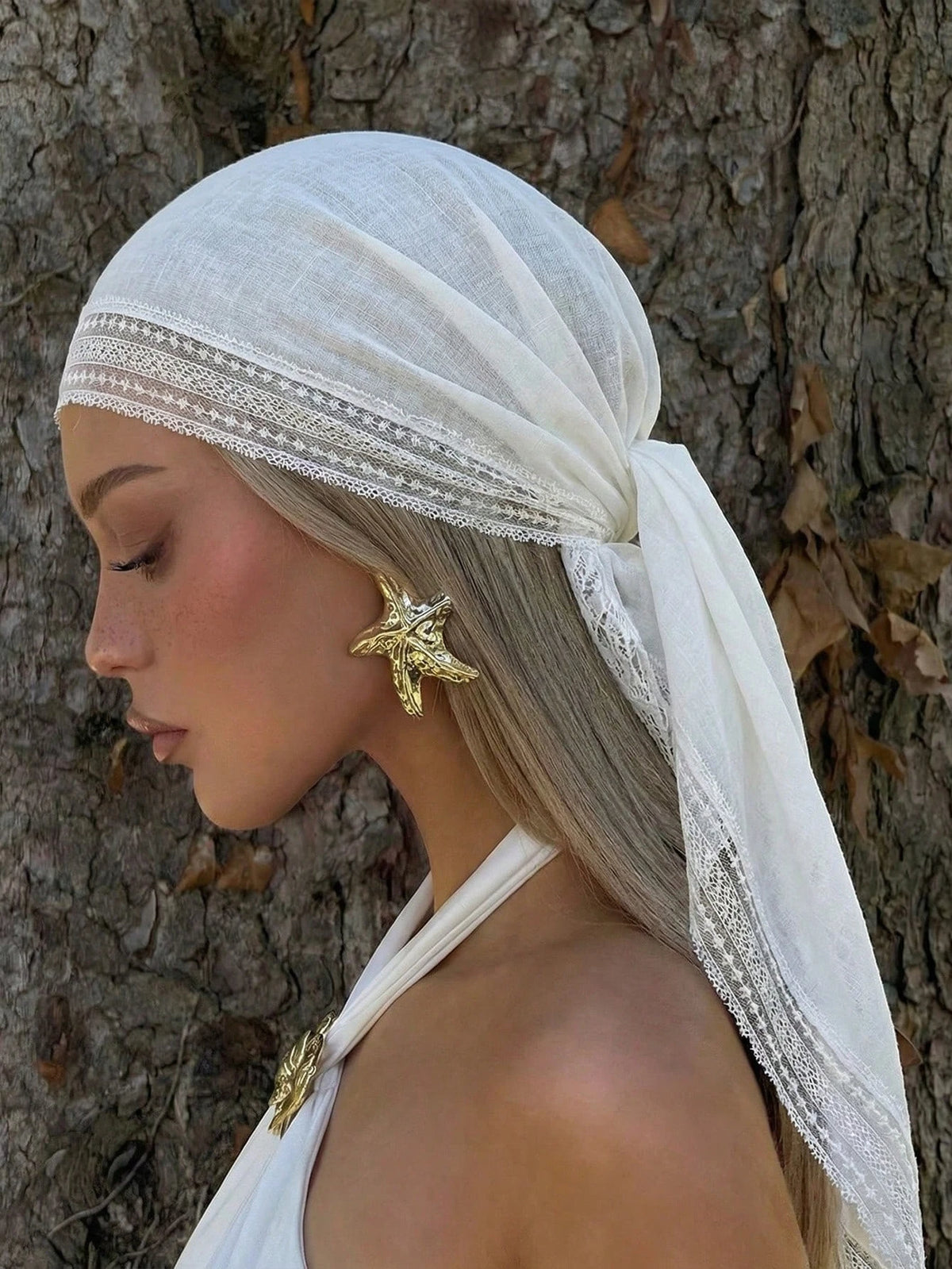 A model wearing a white bohemian lace trim shawl draped over her head and shoulder against a tree trunk.