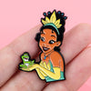 An enamel pin featuring a cartoon woman with black hair and a yellow crown holding a green frog.