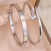 A pair of silver stainless steel bracelets featuring four-leaf clover and nail charms displayed in a light pink box.