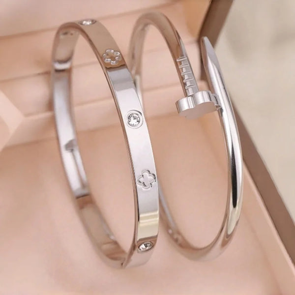 A pair of silver stainless steel bracelets featuring four-leaf clover and nail charms displayed in a light pink box.