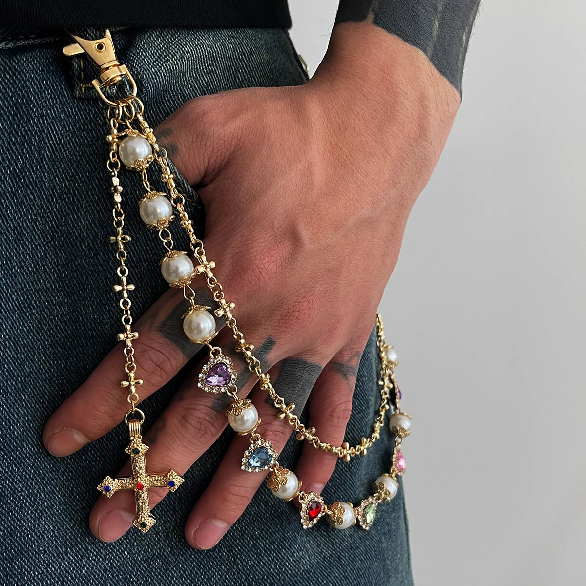 A hand holds a gold pearl cross pendant keychain against a dark gray fabric background.