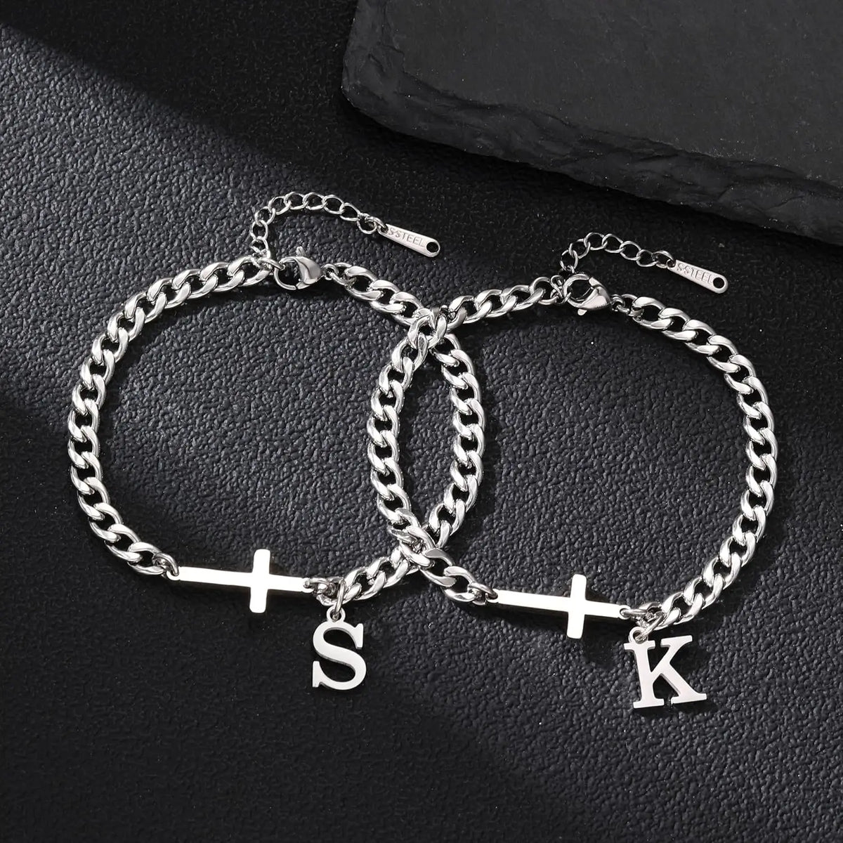 A silver stainless steel Cuban chain bracelet with a cross pendant is displayed on a dark surface.