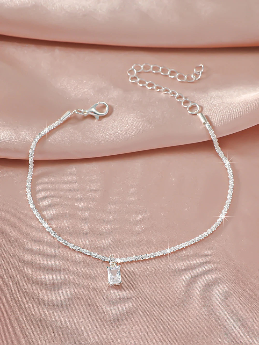 A delicate silver chain anklet with a sparkling zircon heart cross charm displayed on a pink fabric.