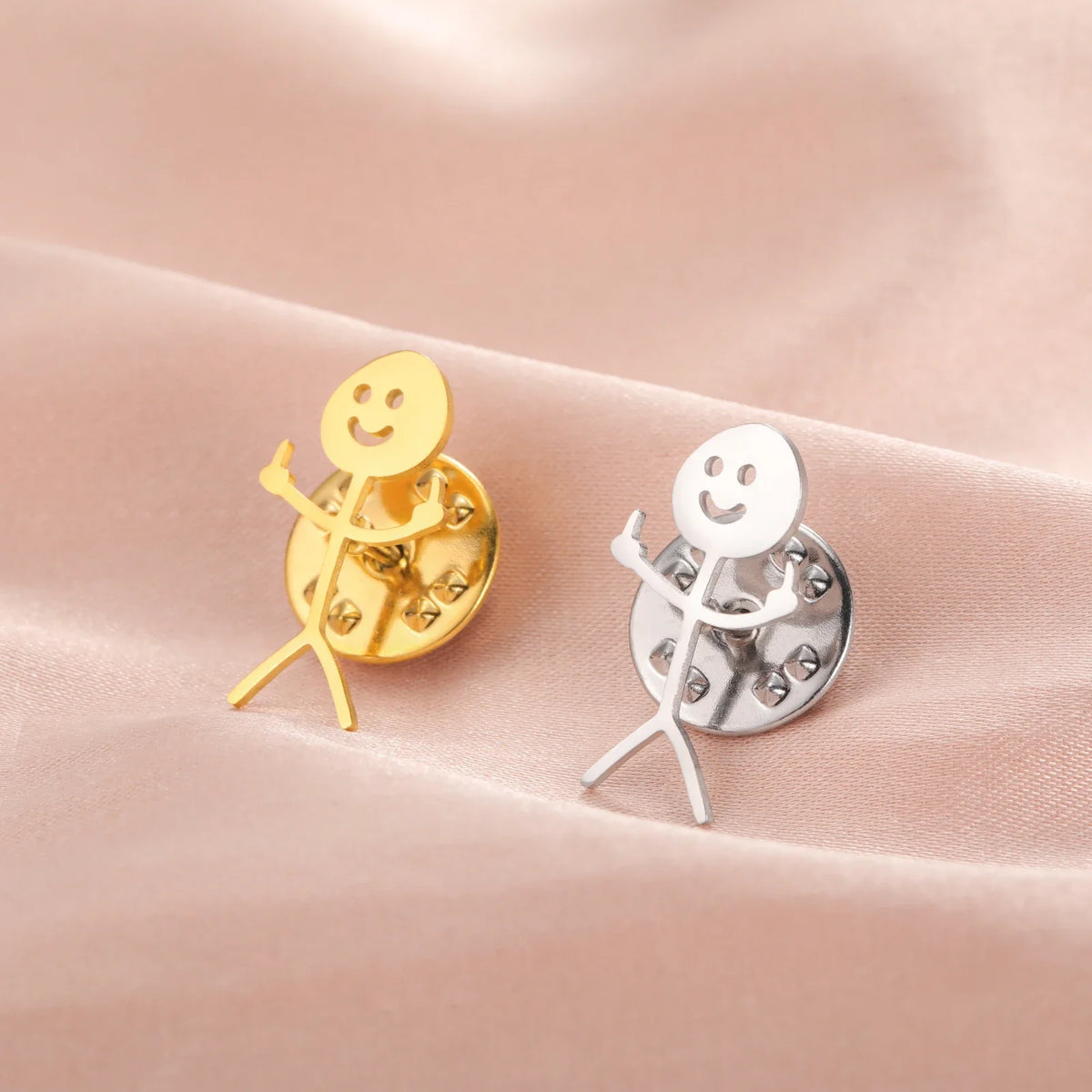 A pair of gold and silver punk stickman brooches resting on a soft pink fabric.