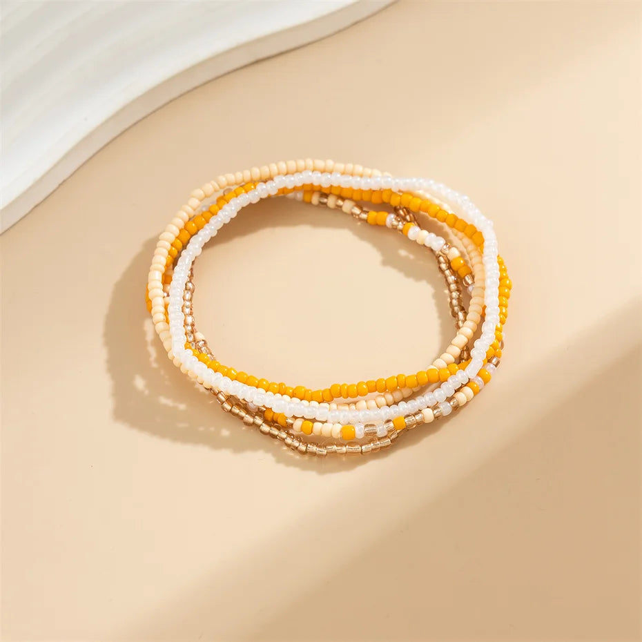 A stack of colorful seed bead anklet bracelets in shades of yellow, white, and orange resting on a beige surface.
