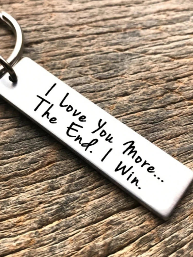 Silver keychain tag engraved with the message I Love You More I Win on a wooden surface.