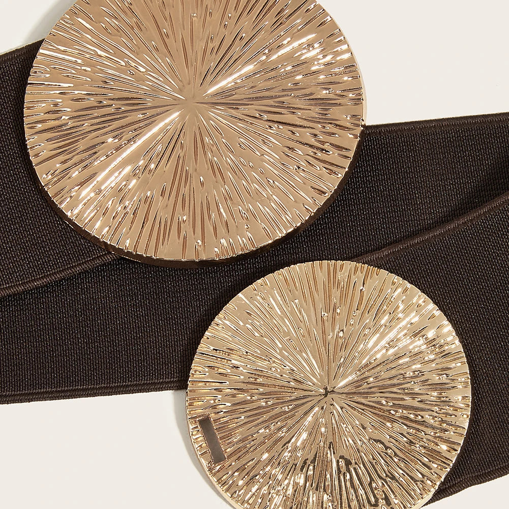 A round, gold-toned metallic belt with a textured, radiating pattern shown against a dark brown fabric background.