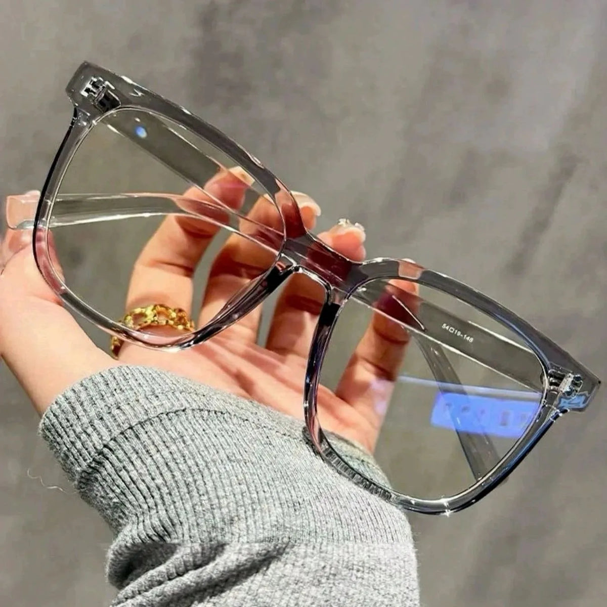 Una mano sostiene unas gafas azules con montura transparente contra un fondo gris.