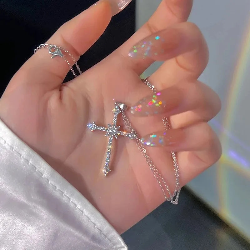 Une main montre un collier de perles minimaliste avec un pendentif croix et des accents de paillettes irisées.