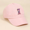 A pink baseball cap featuring a purple and iridescent unicorn letter design on the front.