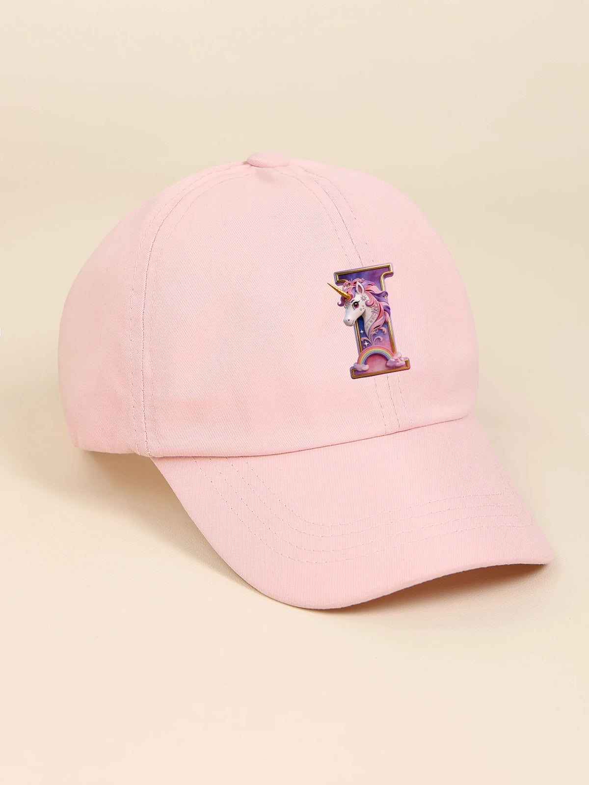 A pink baseball cap featuring a purple and iridescent unicorn letter design on the front.