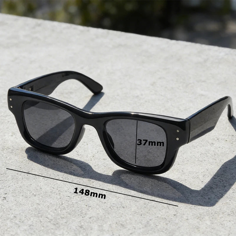 Black square sunglasses resting on a light gray concrete surface with a measurement line.
