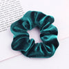A close-up of a dark teal velvet scrunchie with gathered fabric texture lying on white paper.