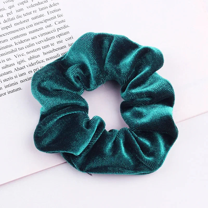A close-up of a dark teal velvet scrunchie with gathered fabric texture lying on white paper.