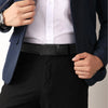 A man wearing a dark blue suit jacket, white shirt, and black trousers, holding a black leather ratchet dress belt.
