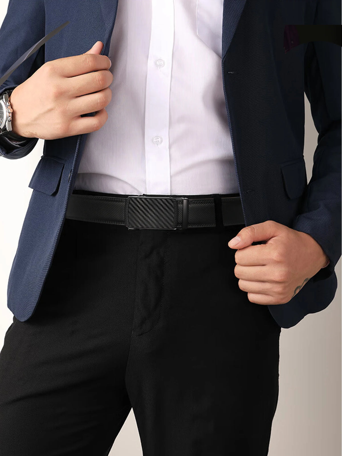 A man wearing a dark blue suit jacket, white shirt, and black trousers, holding a black leather ratchet dress belt.