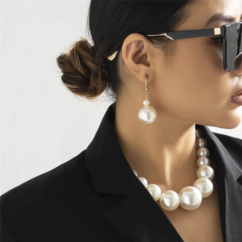 Une femme portant des lunettes de soleil noires et un blazer noir avec un élégant collier imitation perles.