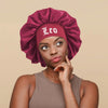 A woman wearing a deep magenta satin sleep bonnet with a Leo wrap band and a simple brown tank top.