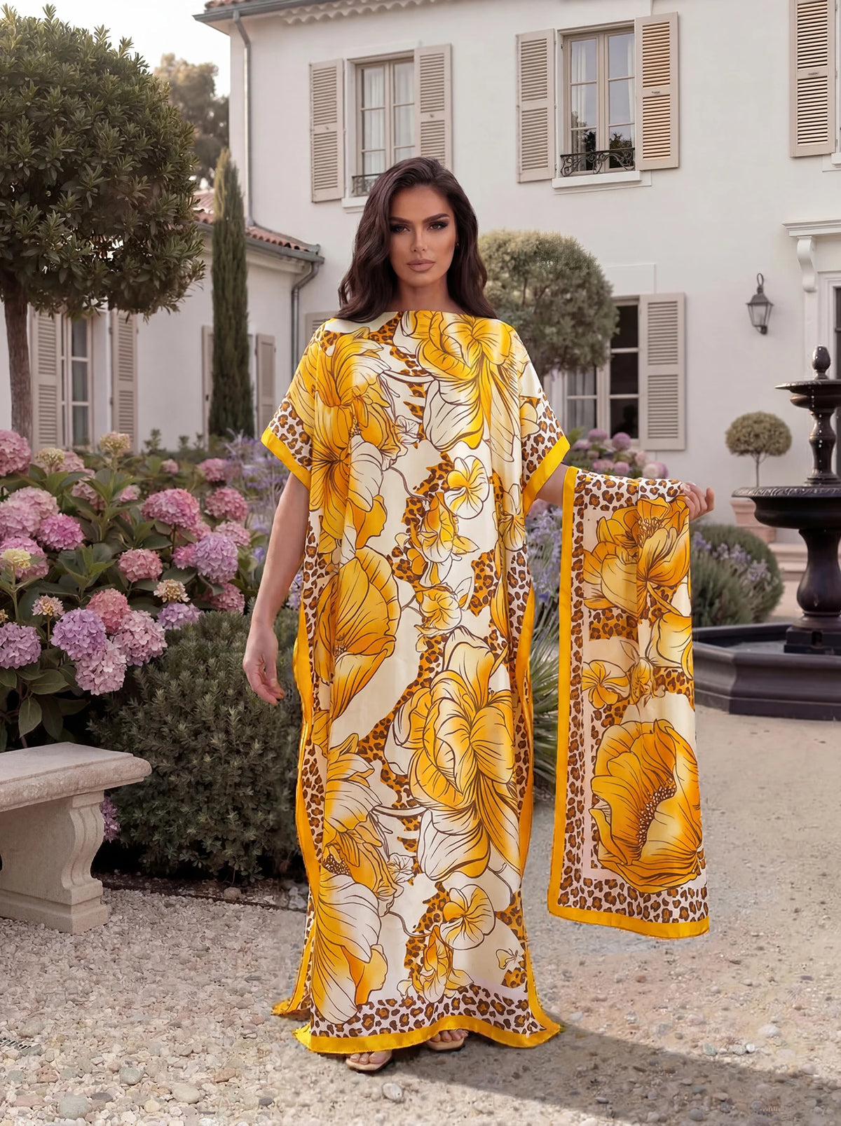 A woman models a yellow and brown tropical floral leopard print abaya scarf outdoors in front of a white building.