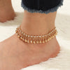 A gold bell foot chain anklet worn on a bare foot against a white fluffy background.