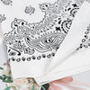 A close-up of a white cotton bandana scarf featuring a black paisley pattern, displayed folded with floral greenery.