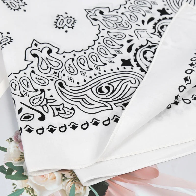 A close-up of a white cotton bandana scarf featuring a black paisley pattern, displayed folded with floral greenery.