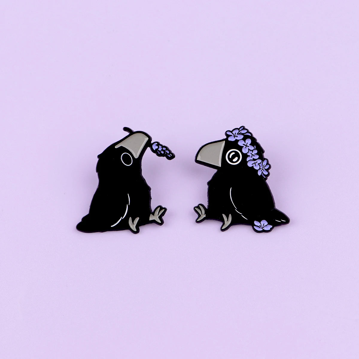 Ein Paar schwarze Email-Anstecker in Form von Krähen mit lila Blumen auf dem Kopf.