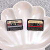 A pair of retro cassette enamel pins labeled MIX TAPE and AWESOME MIX VOL placed on a white seashell dish.