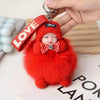A bright red fluffy plush doll with a bow and a leather keychain labeled LOVE is being held by a hand.