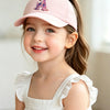 A smiling young girl wearing a pink rainbow unicorn letter baseball cap and a white dress.