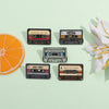Collection de pins en émail de cassettes vintage et une fleur blanche sur fond vert clair.