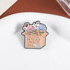 An enamel flower brooch featuring pink and blue wildflowers growing out of a box with the text Consider how the wild flowers 
