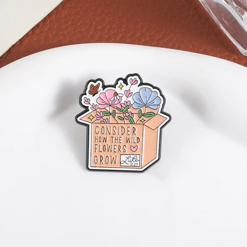 An enamel flower brooch featuring pink and blue wildflowers growing out of a box with the text Consider how the wild flowers 